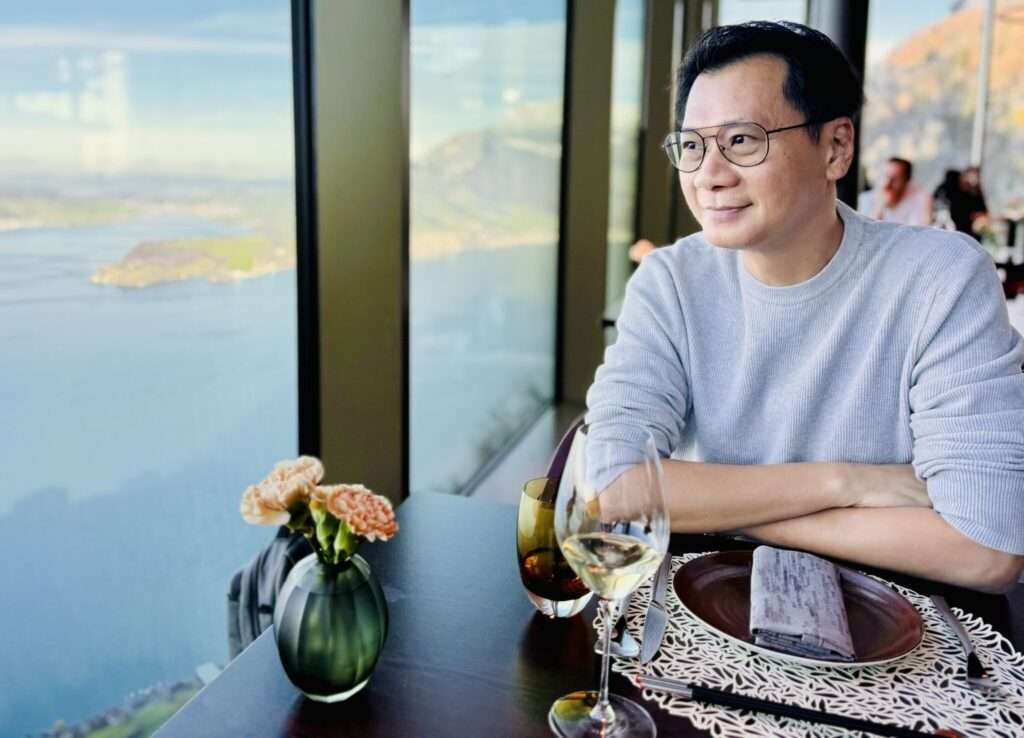 Krish Phua dining at a restaurant with panoramic views of lake Lucerne, with a glass of white wine and flowers on the table